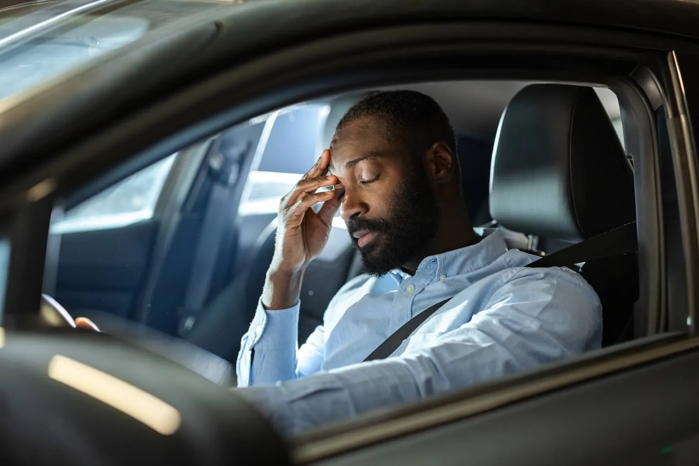 Tired man sitting in the driver’s seat of a car with closed eyes, one hand on the steering wheel and the other hand raised to the head.