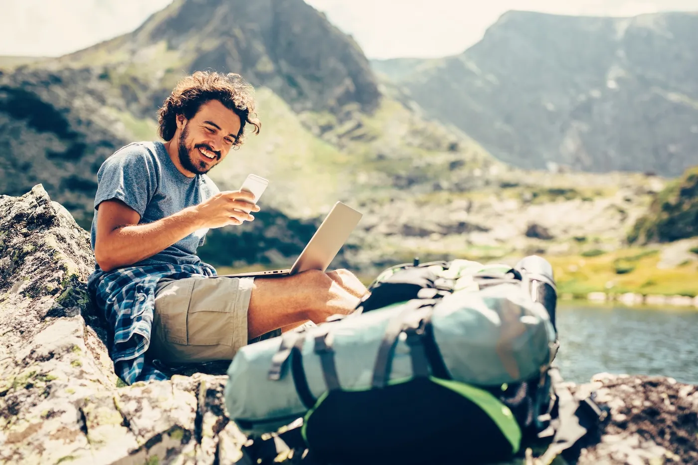 Male hiker resting and using his smart phone on a rock near a lake in the mountain.