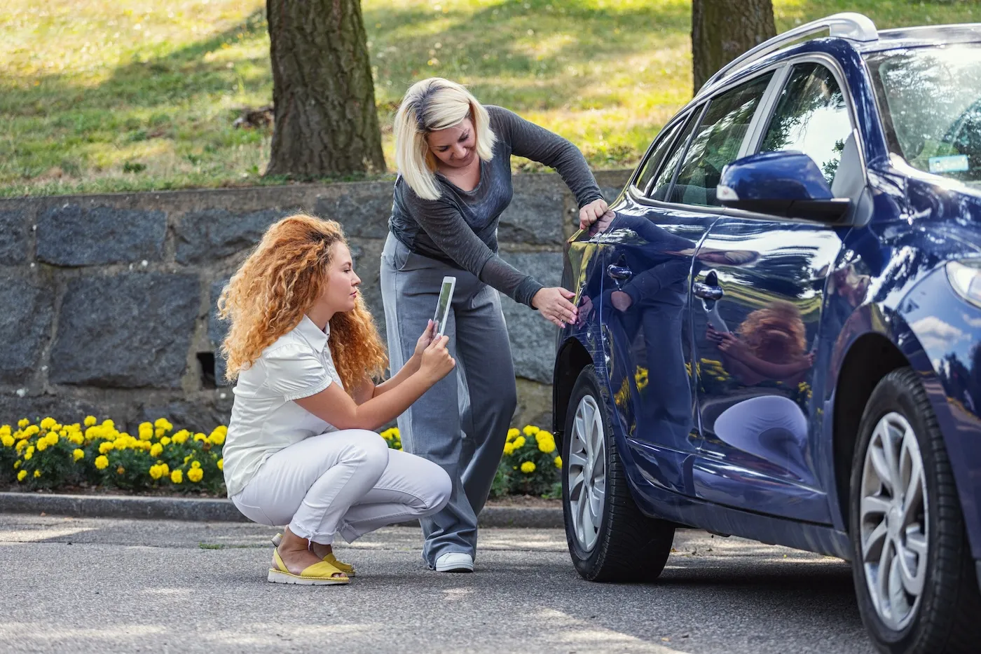 Two women examining a dent on the back of a blue car. One is taking a photo of the damage.