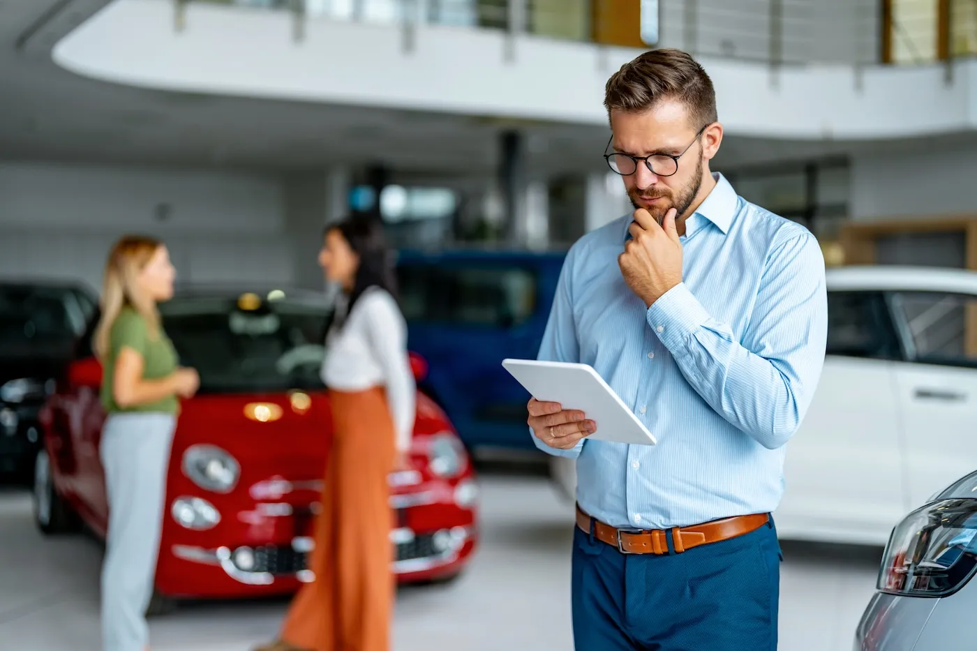 Serious salesman using digital tablet in a car showroom. Two women are talking next to a red car in the background.