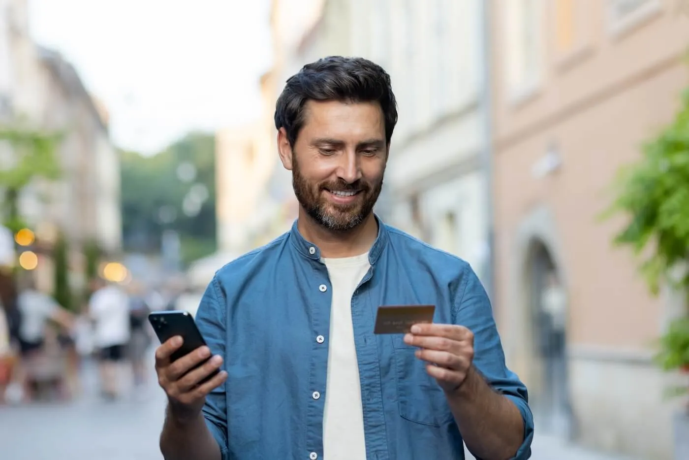 Smiling man in the street looking at his credit card and holding a smartphone