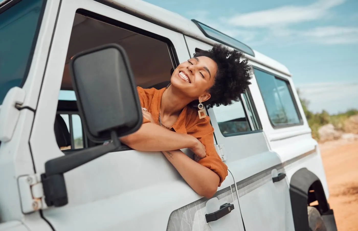 Smiling woman leans out of the open window of a white off‑road vehicle on a sunny day, resting her arms on the door while looking toward the landscape.