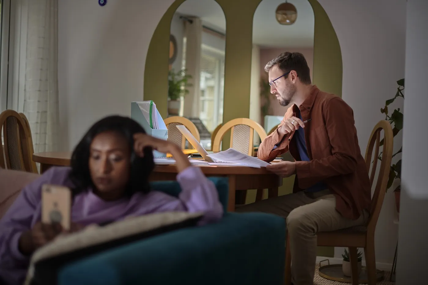 A man poring over documents at a table late at night. His friend is sitting on the couch on her phone.