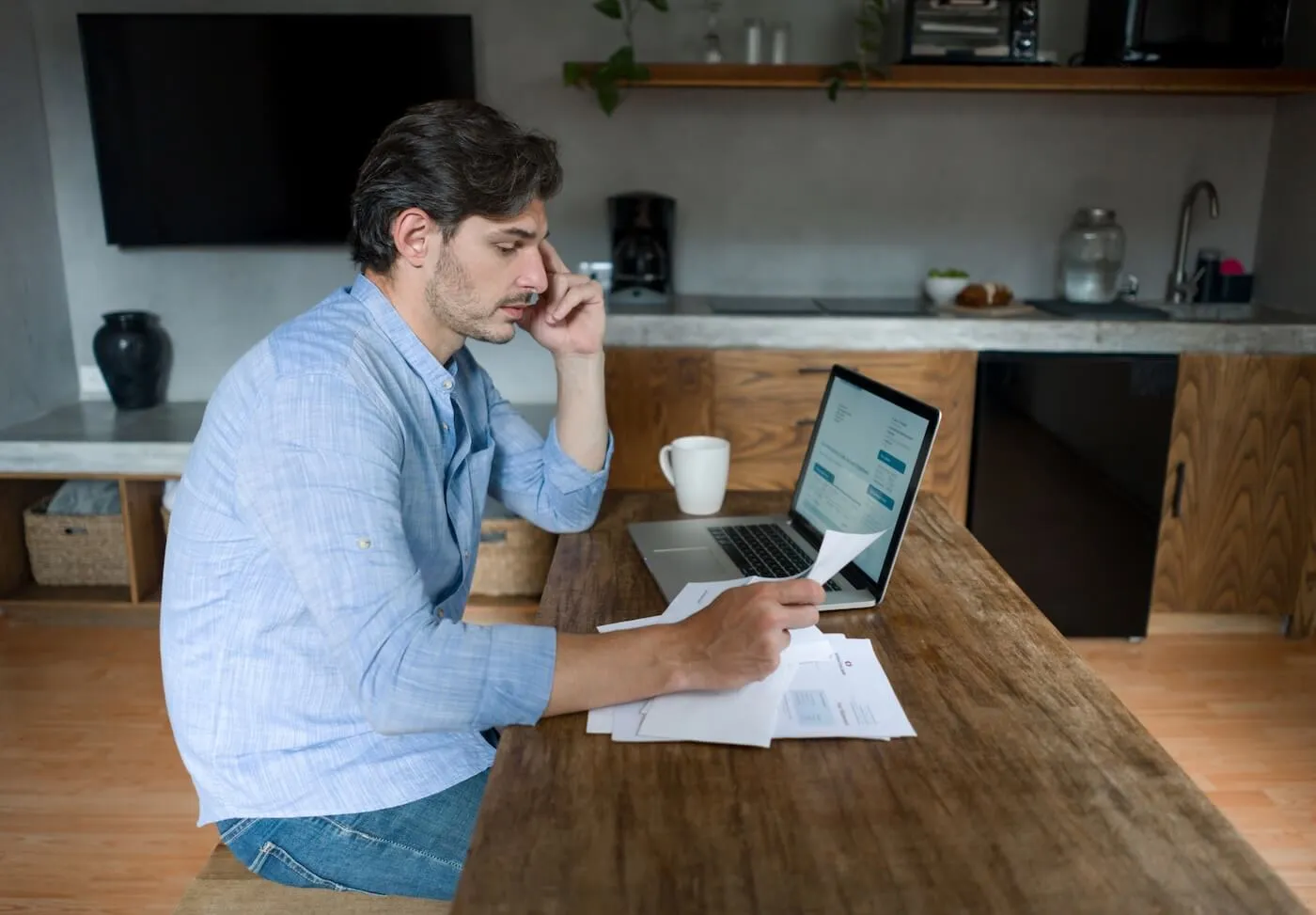 Concerned man reviewing paperwork at a kitchen table while working on a laptop at home