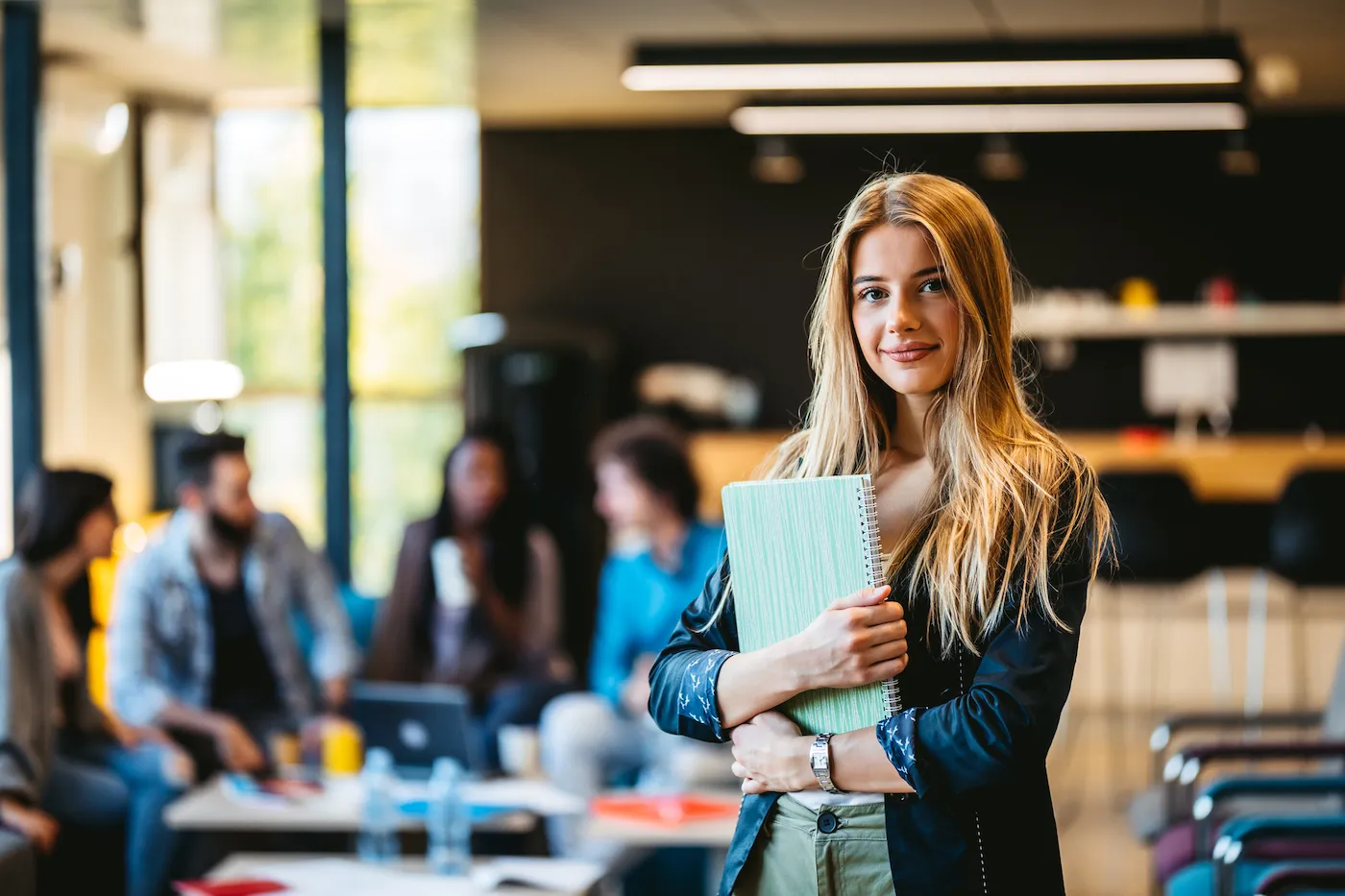 Student in library holding notebook, with student peers studying at a table in the background.