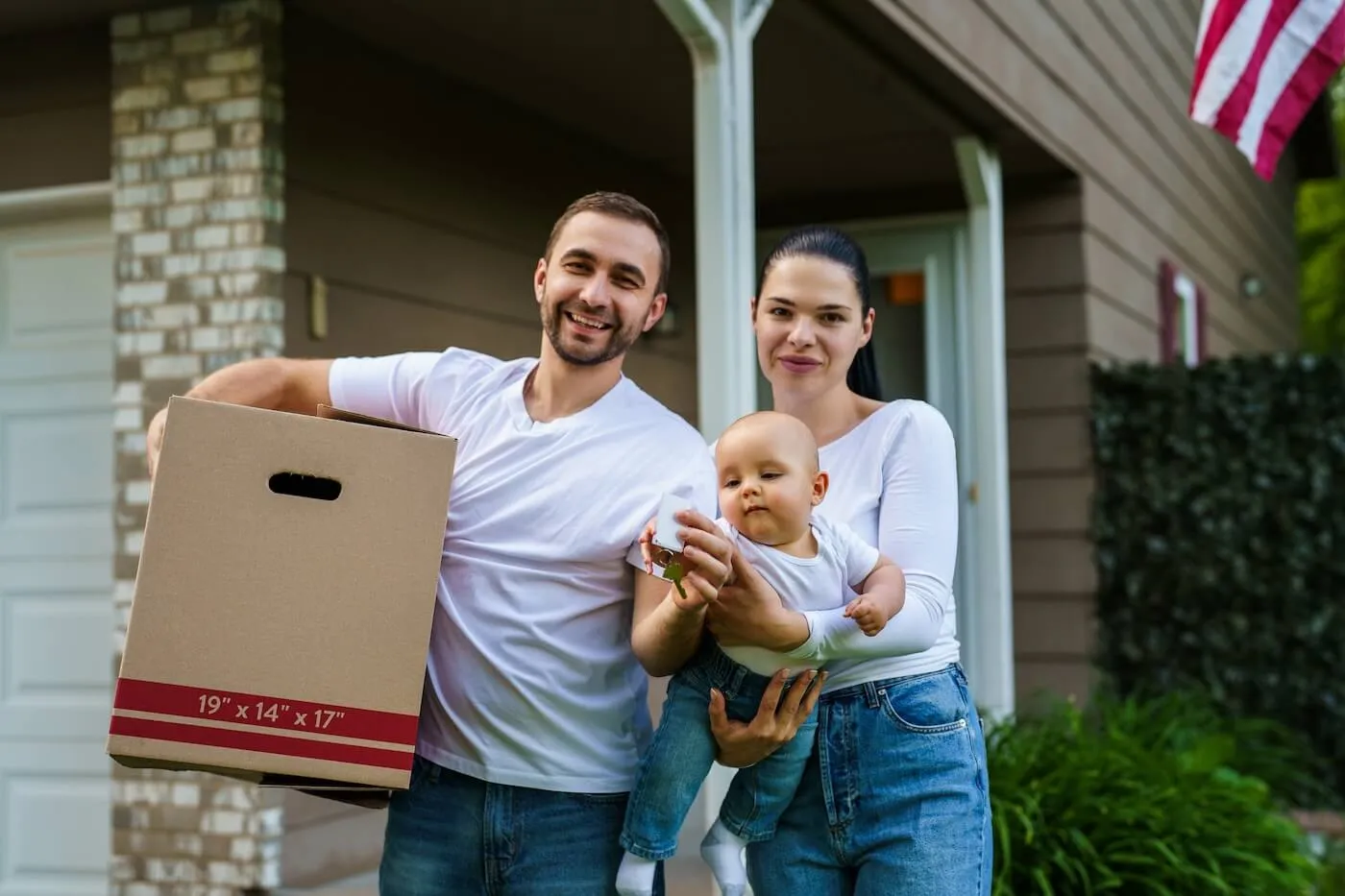 Happy family with a baby standing in front of their new house, the man is holding a moving box and the house keys