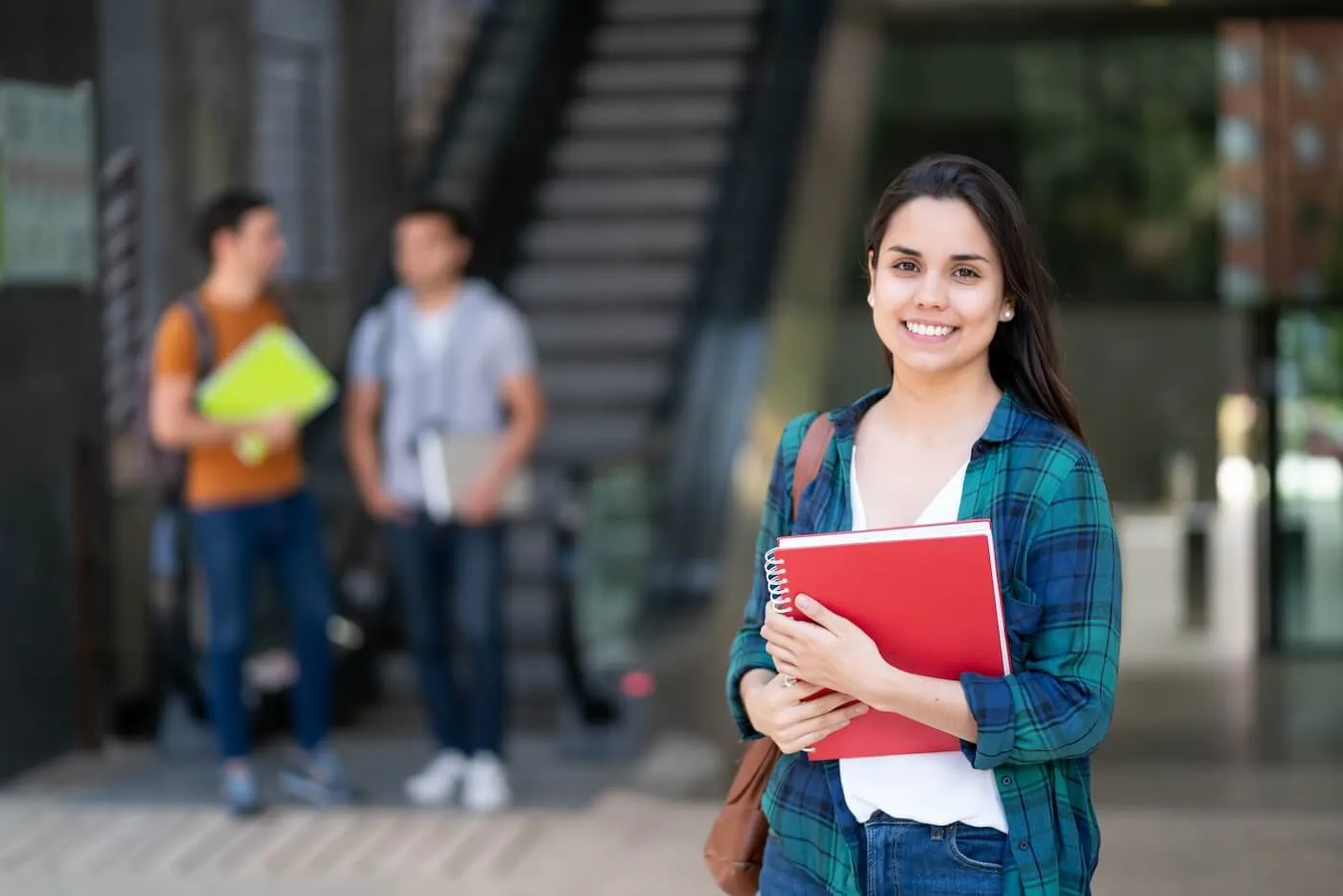 Smiling female student is holding her books while posing for photo, two male student are chatting on the background next to an escalator