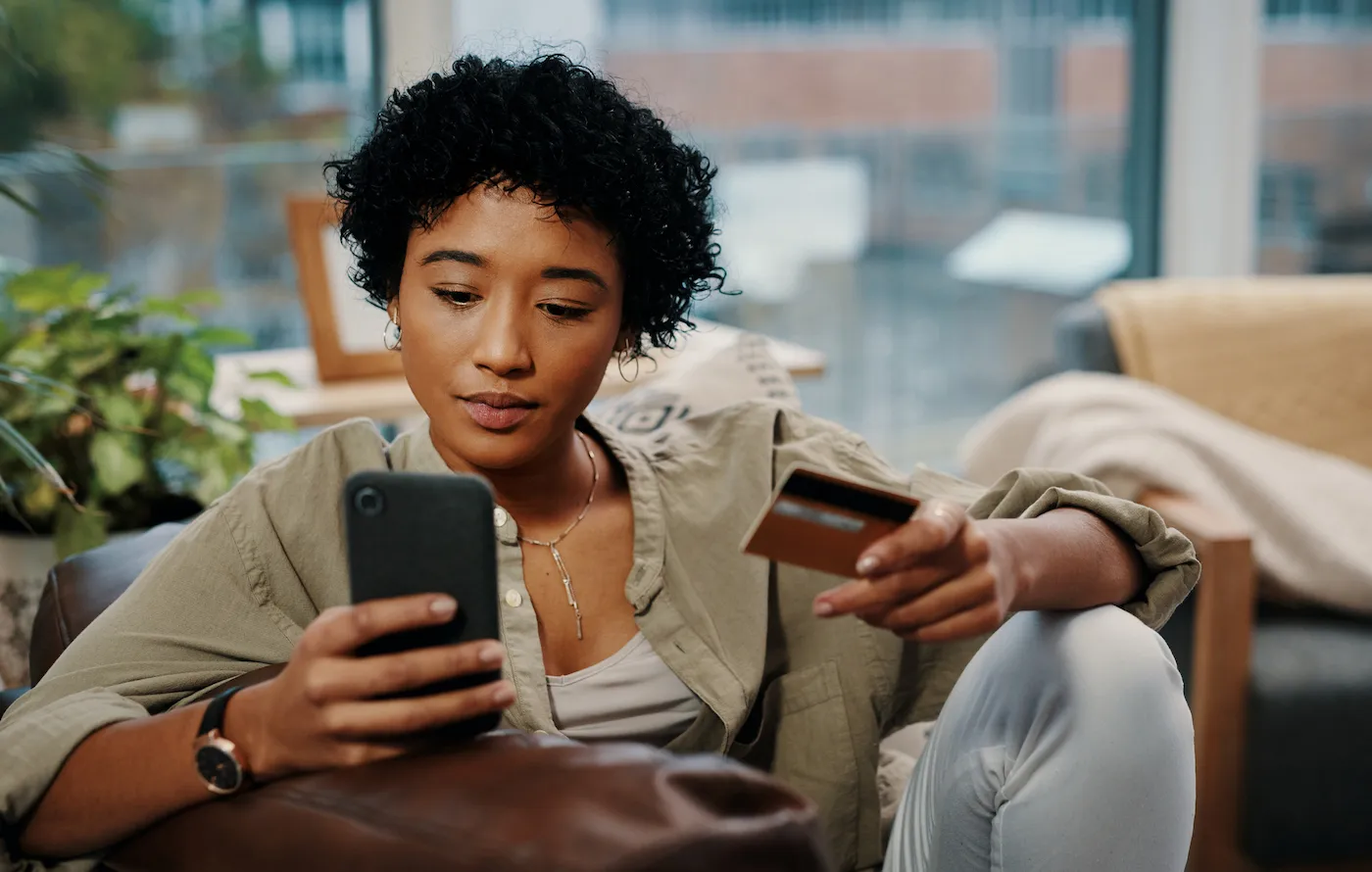 Shot of a young woman holding her credit card while using her cellphone to apply for credit card preapproval at home