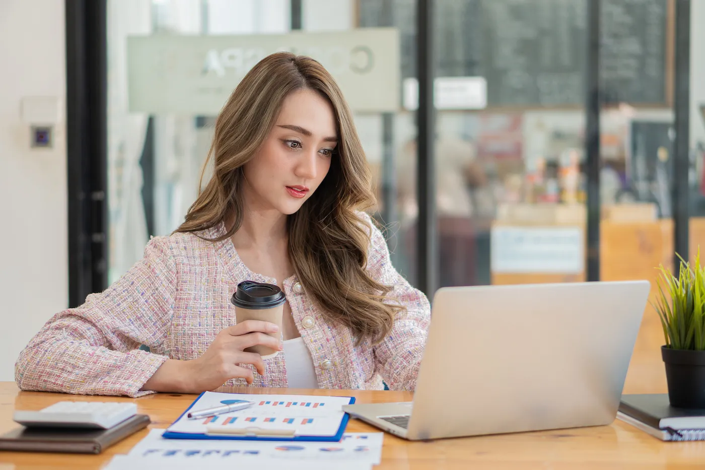 A woman avoiding emotional investing, holding a coffee cup and looking at a laptop while sitting at a desk with papers.