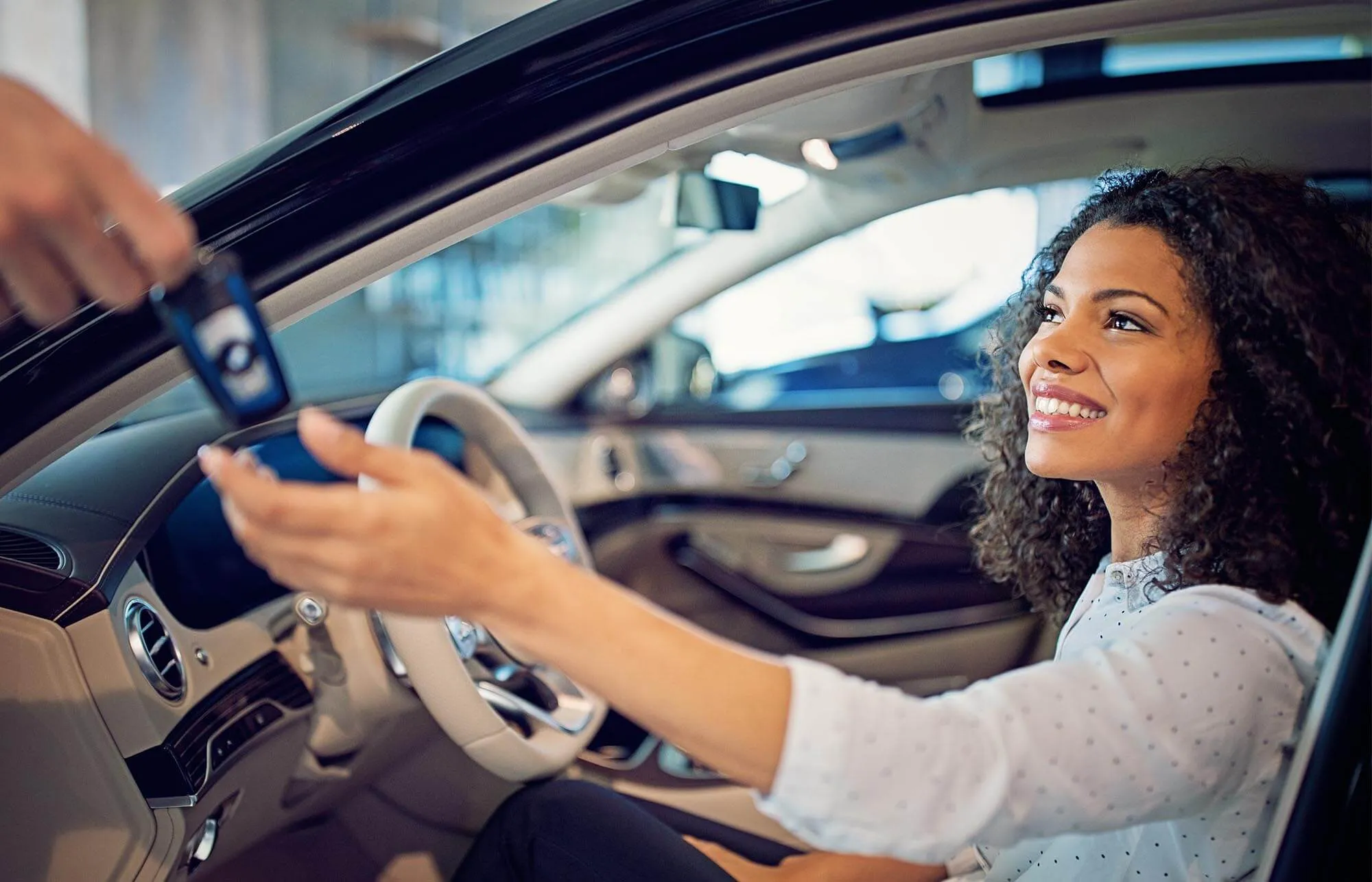 A woman being handed car keys while sitting in the driver's seat of a car parked in a dealership.