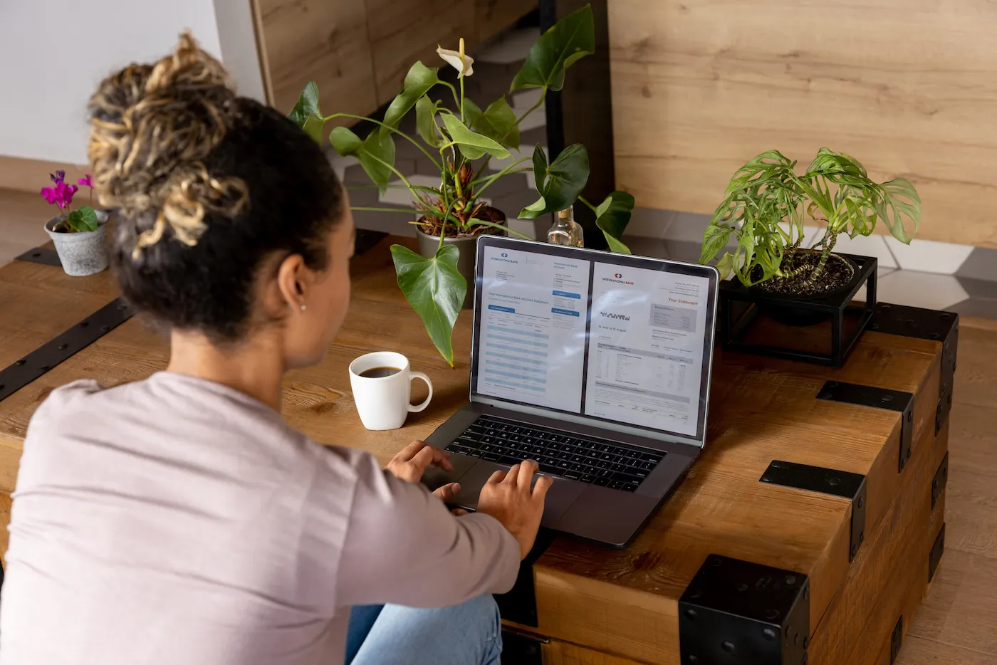 Woman at home checking her rewards checking account statements online using a laptop computer on a wooden trunk coffee table with plants on it.