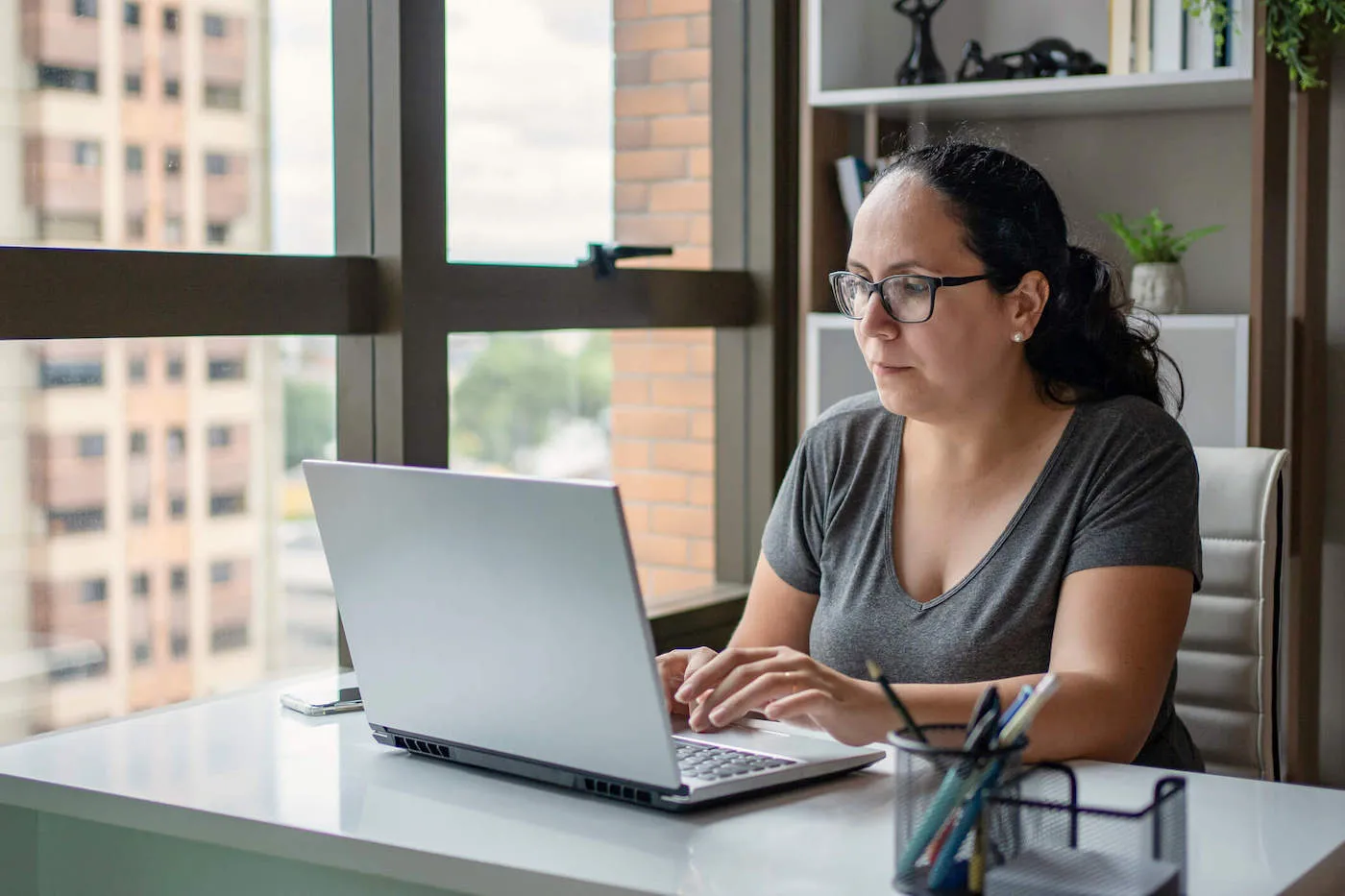 Woman in glasses typing on laptop while seated by window.