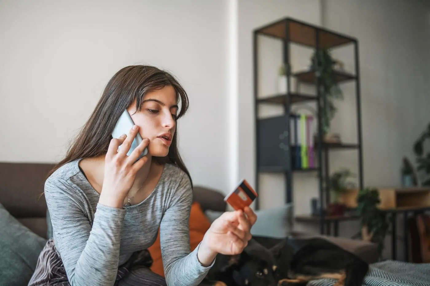 Focused woman seated on a sofa in a living room, holding a credit card while speaking on a smartphone. Shelving, houseplants, and home furnishings are visible in the background.