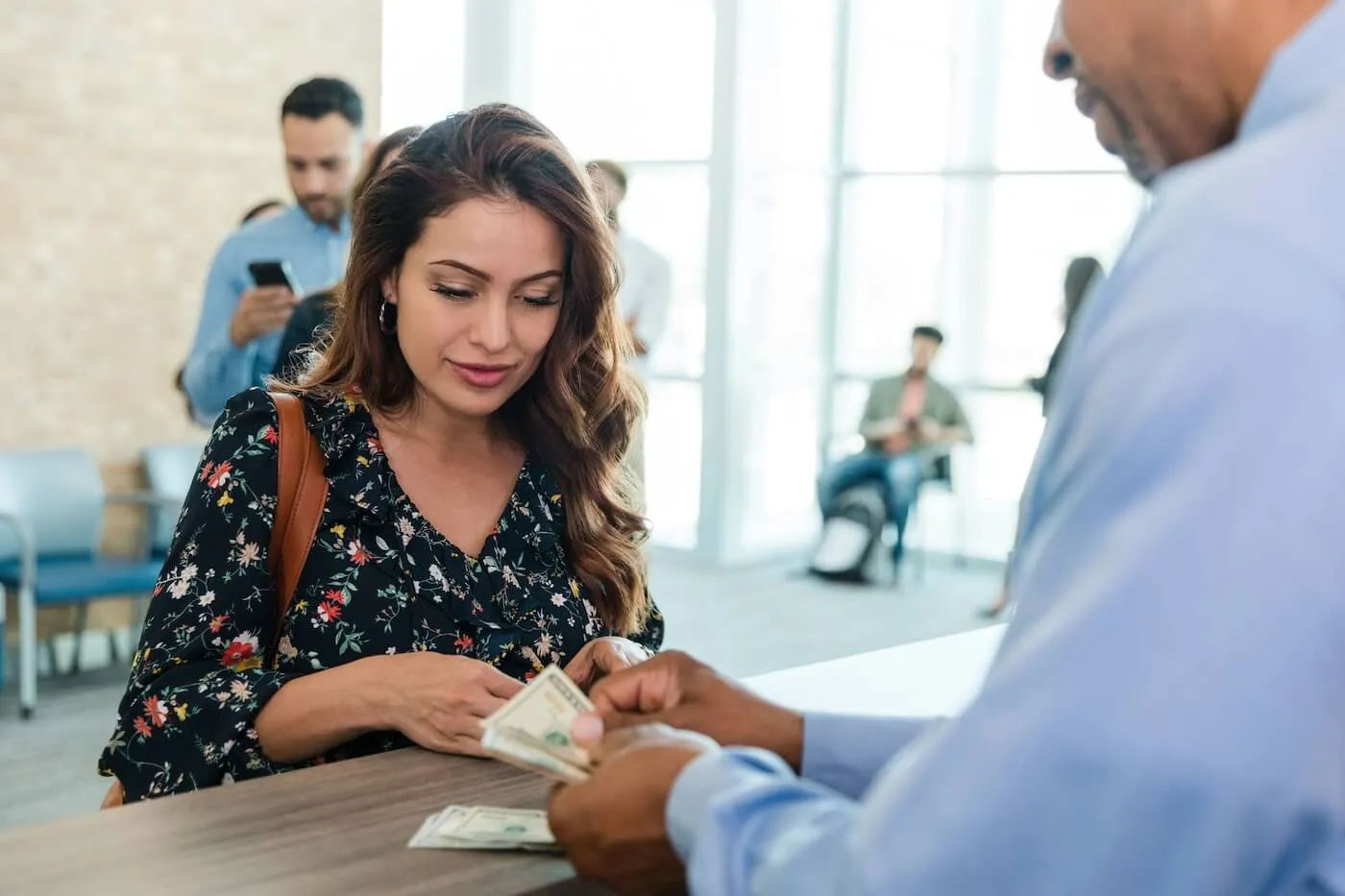 Woman standing at a customer service counter while a bank employee counts and hands over cash. Other customers are visible in the background of a bright financial institution