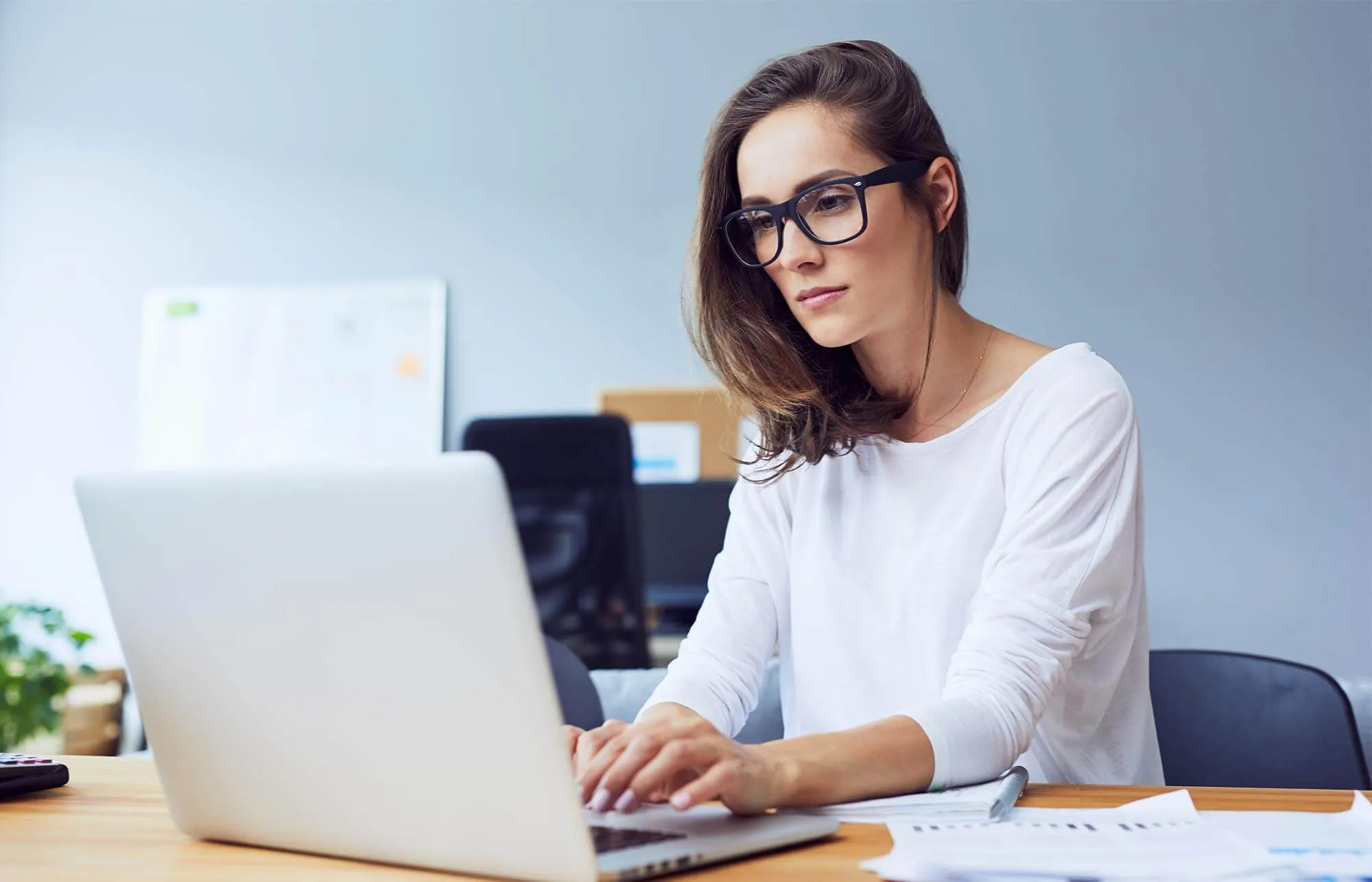 A woman wearing glasses and white shirt is working on the laptop at her desk