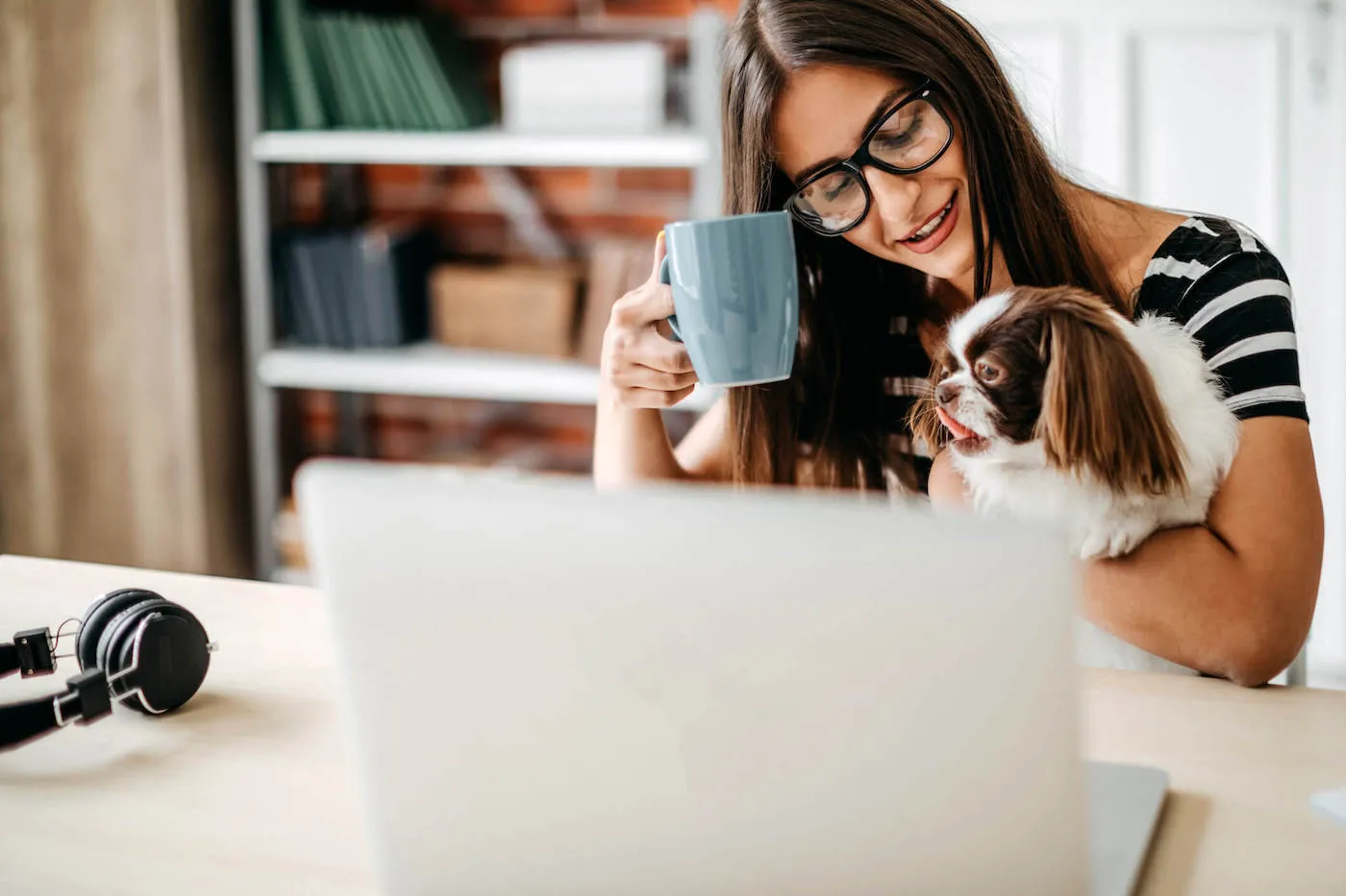 Young woman in glasses holding a cup and pet dog while seated in front of open laptop screen.