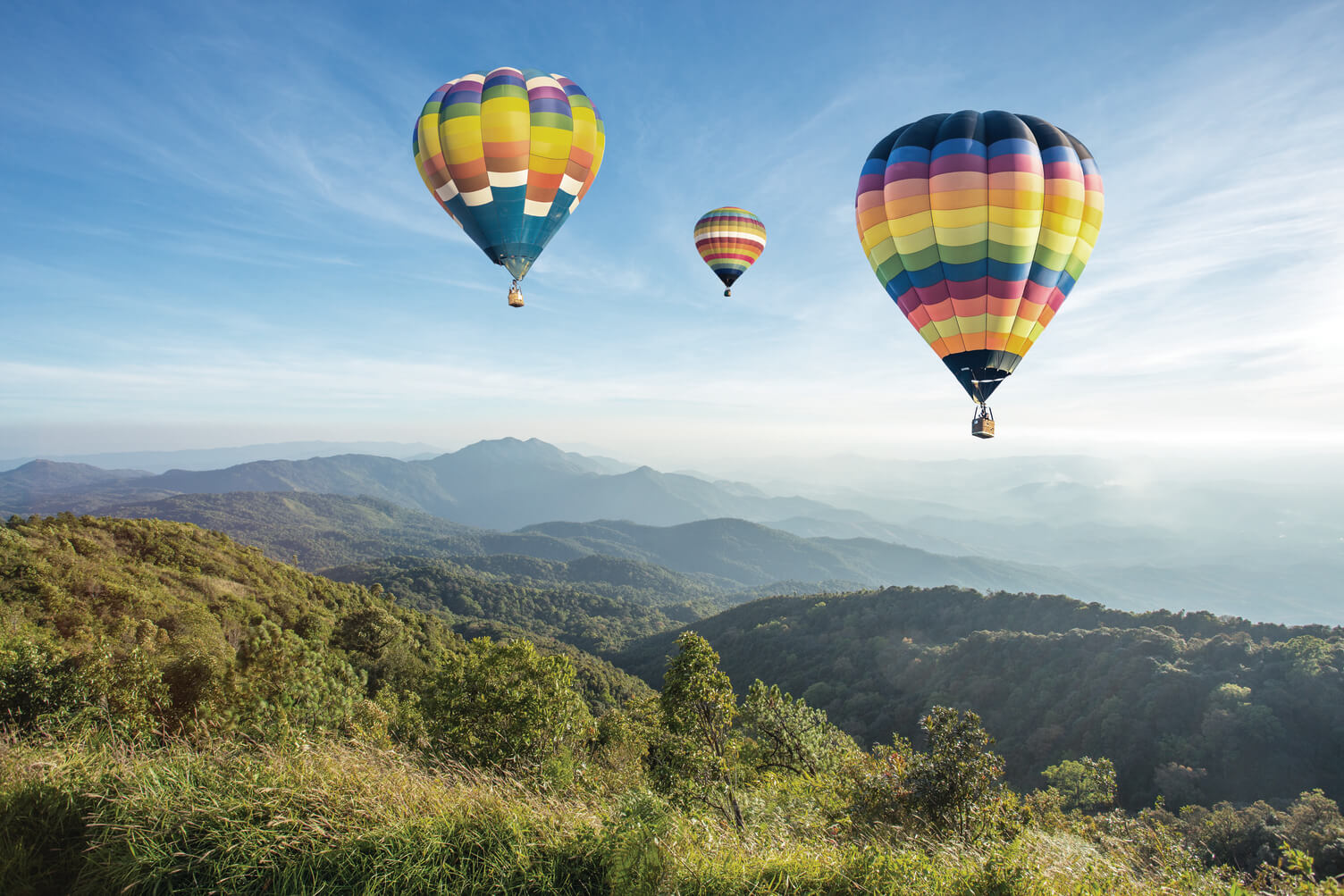 Three colorful hot air balloons floating in a clear blue sky. The balloons are yellow, blue, green, and red, each with unique patterns. They are soaring above a scenic landscape with green fields and distant mountains.