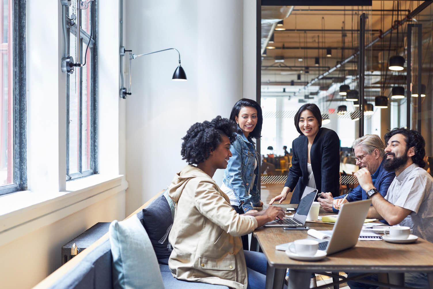 Diverse group of adults collaborate at a long table in a modern office, using laptops and documents in a bright, open workspace.