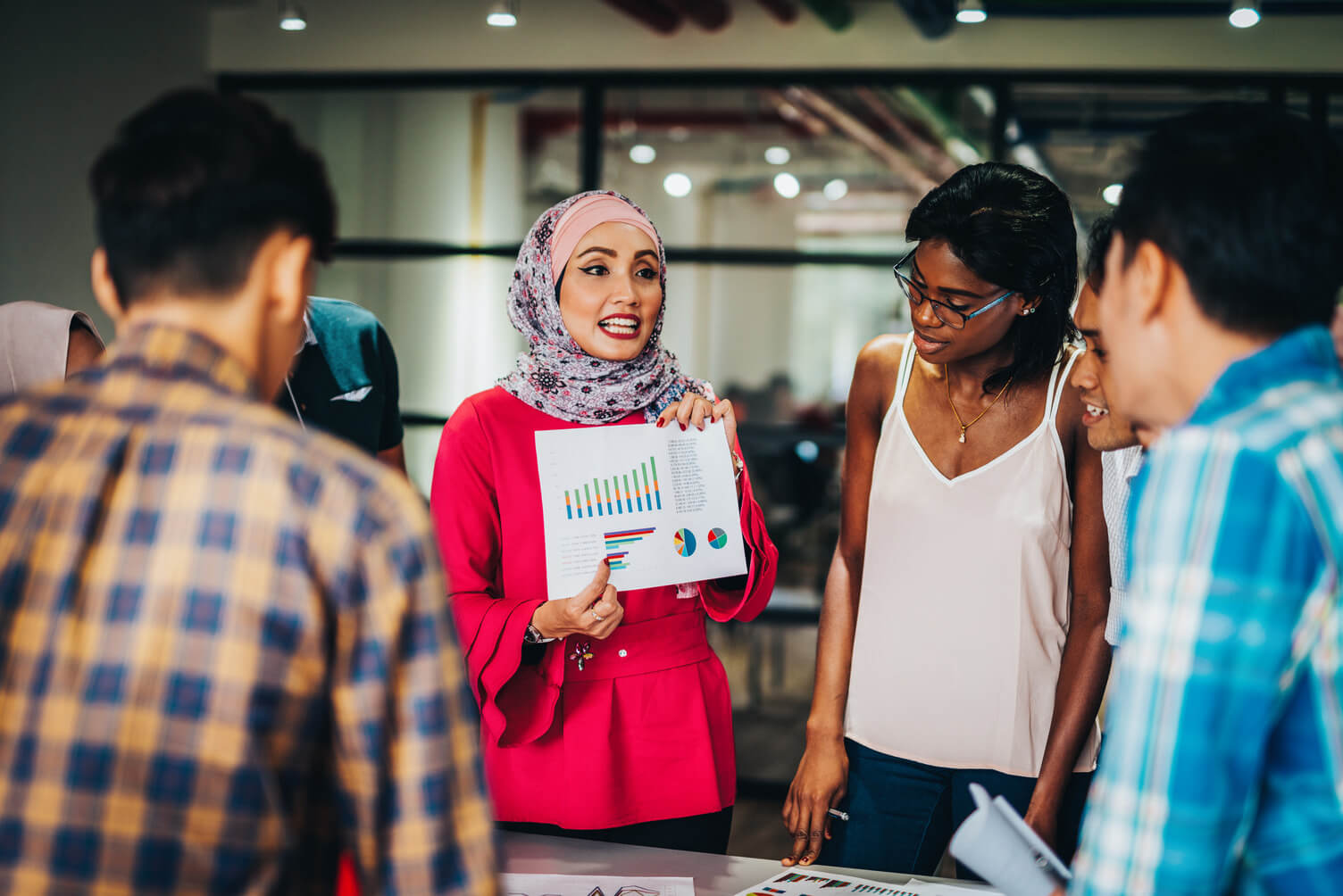 Compliance and accuracy: A group of people standing around a table in a modern office setting. One person is holding a sheet with colorful charts and graphs, presenting information to the group. Other papers are spread out on the table.
