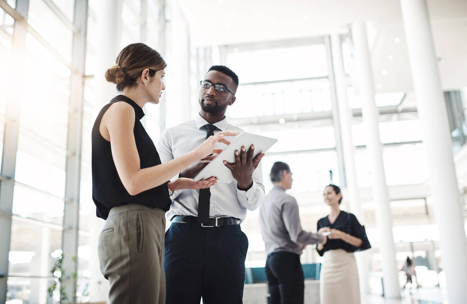 Two people standing in a bright, modern office space, looking at a tablet together while others talk in the background.