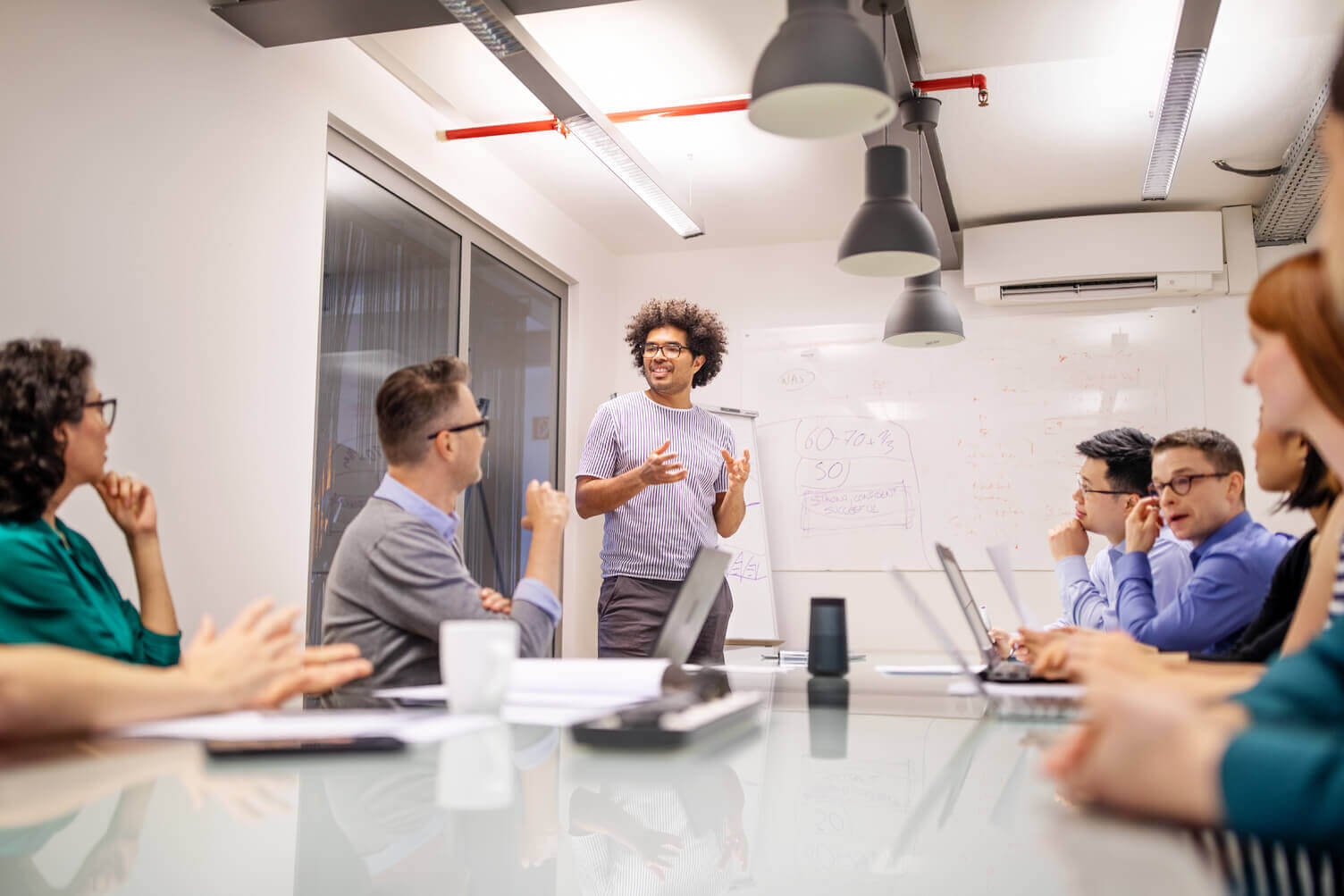 Take the burden out of tax management: A person standing and speaking to a group in a modern conference room. Several people are seated around a table with laptops and papers, while a whiteboard with diagrams is visible in the background.