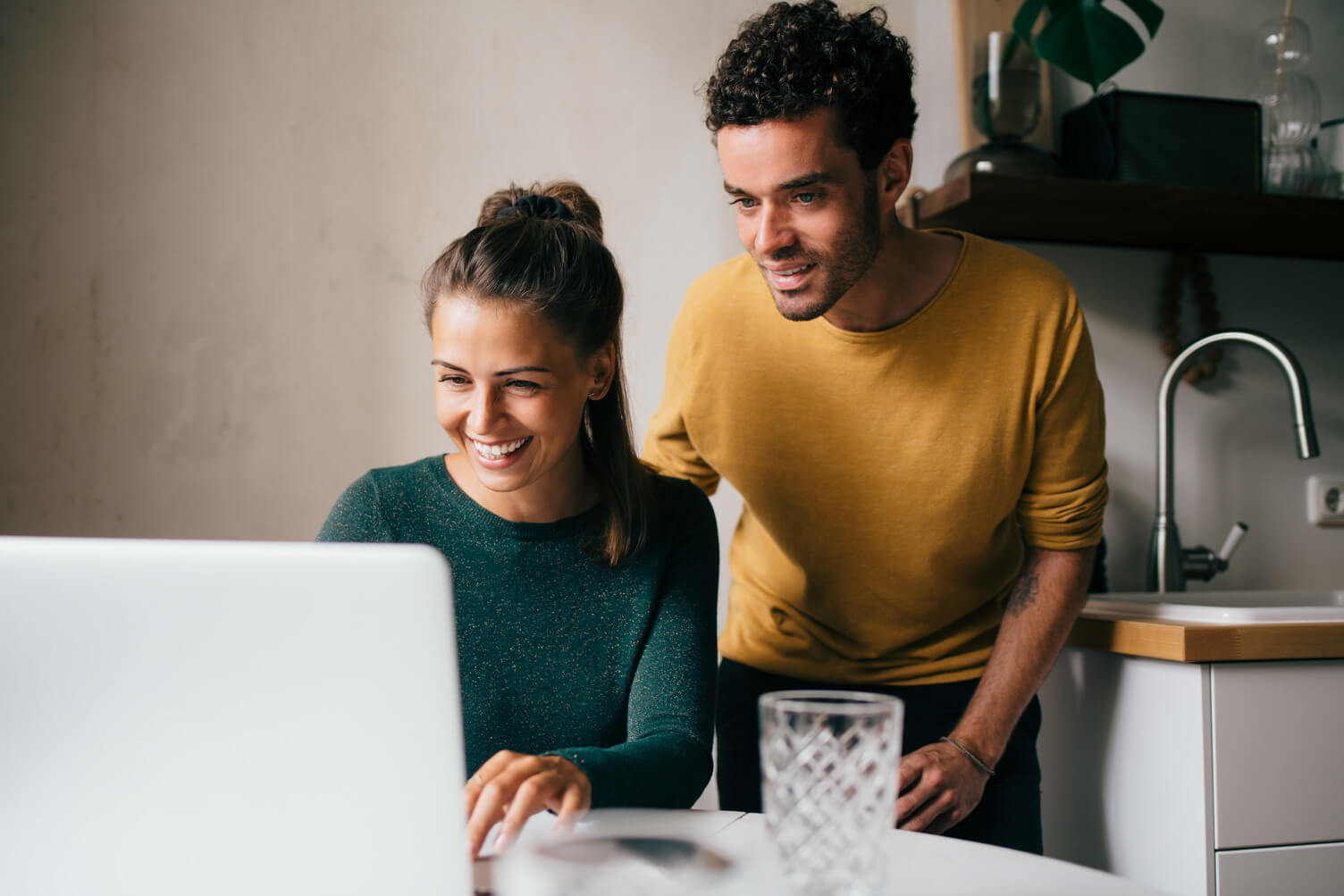 Two adults work together at a kitchen table, reviewing information on a laptop in a home workspace, with shelves and a sink in the background, representing remote work and digital employment verification through Experian Verify™ employer services.