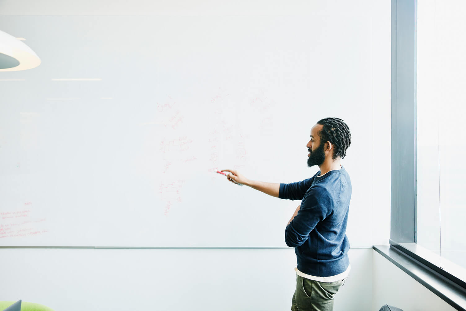 A Black man with short braided hair and a beard stands in a bright, modern office, facing a large whiteboard. He wears a long-sleeve sweater and casual work pants and gestures toward faint handwritten notes on the board. Natural light comes through tall windows, suggesting a collaborative workplace setting focused on planning, workforce management, or problem-solving.