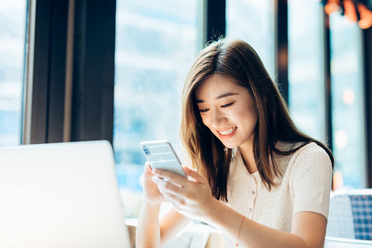 Asian woman in her 20s–30s sits at a desk in a bright modern office, smiling while using a smartphone beside an open laptop, suggesting mobile access to employer services, workforce management, or hiring tools.