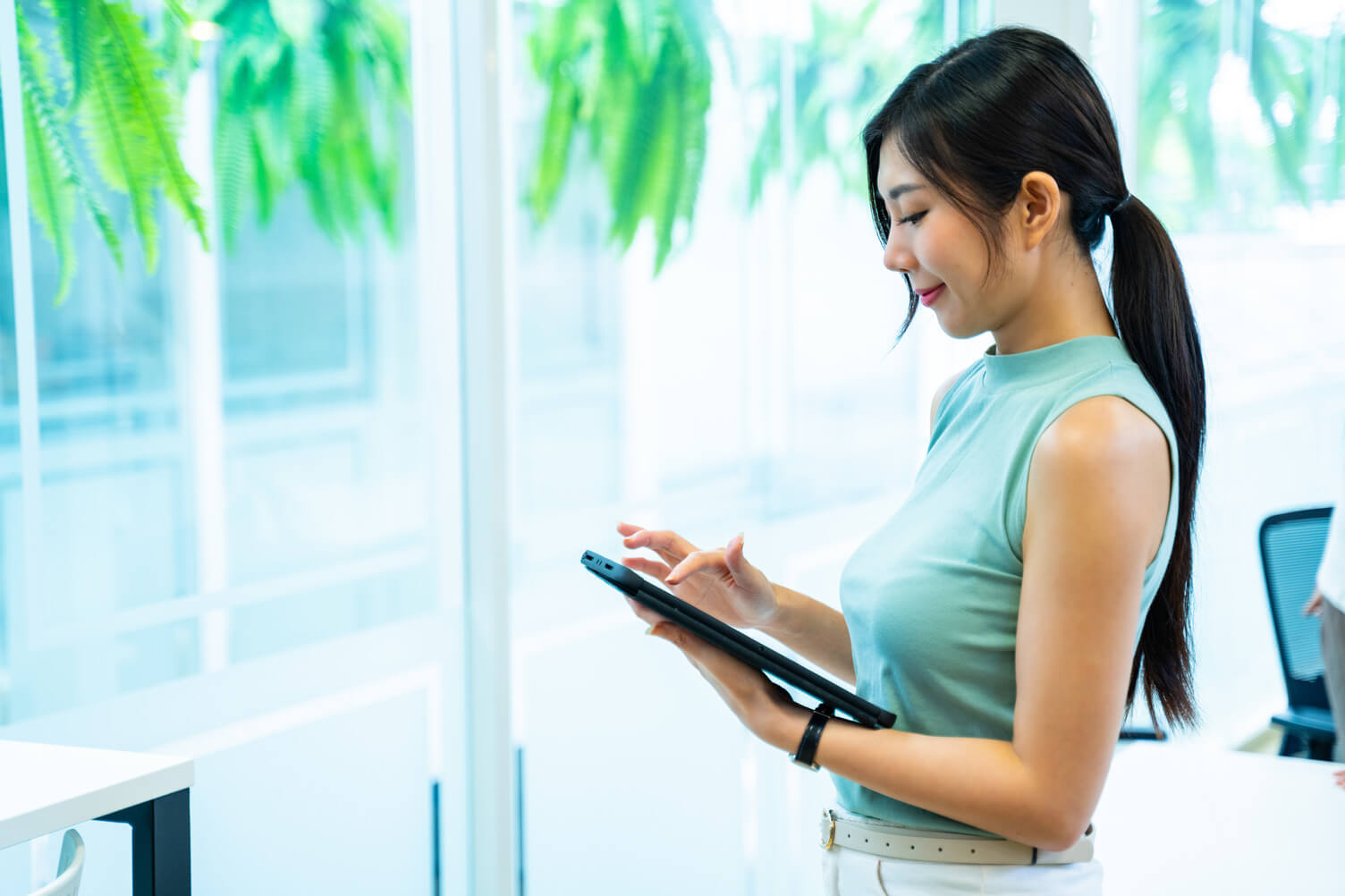 Person standing in a bright office holding a tablet, with green hanging plants near large windows