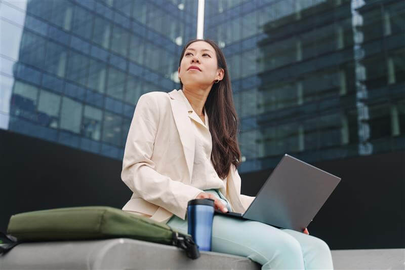 An Asian woman in a beige blazer sits outdoors with a laptop and travel mug in front of a modern glass office building.