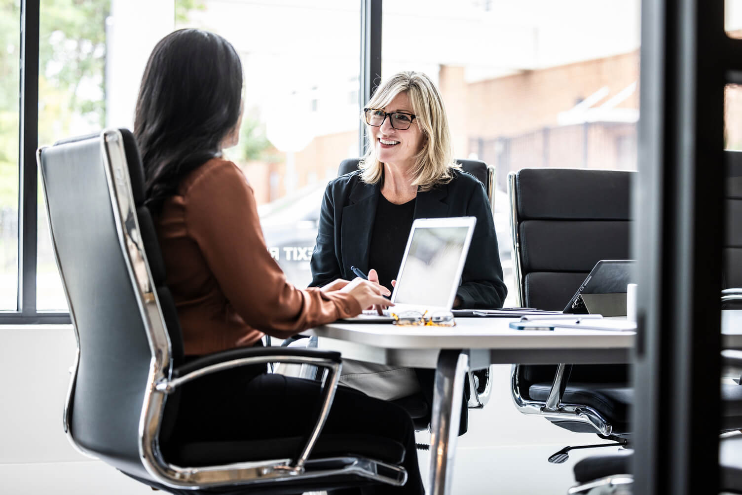 Two professionals seated at a modern office table with laptops, engaged in a business meeting.