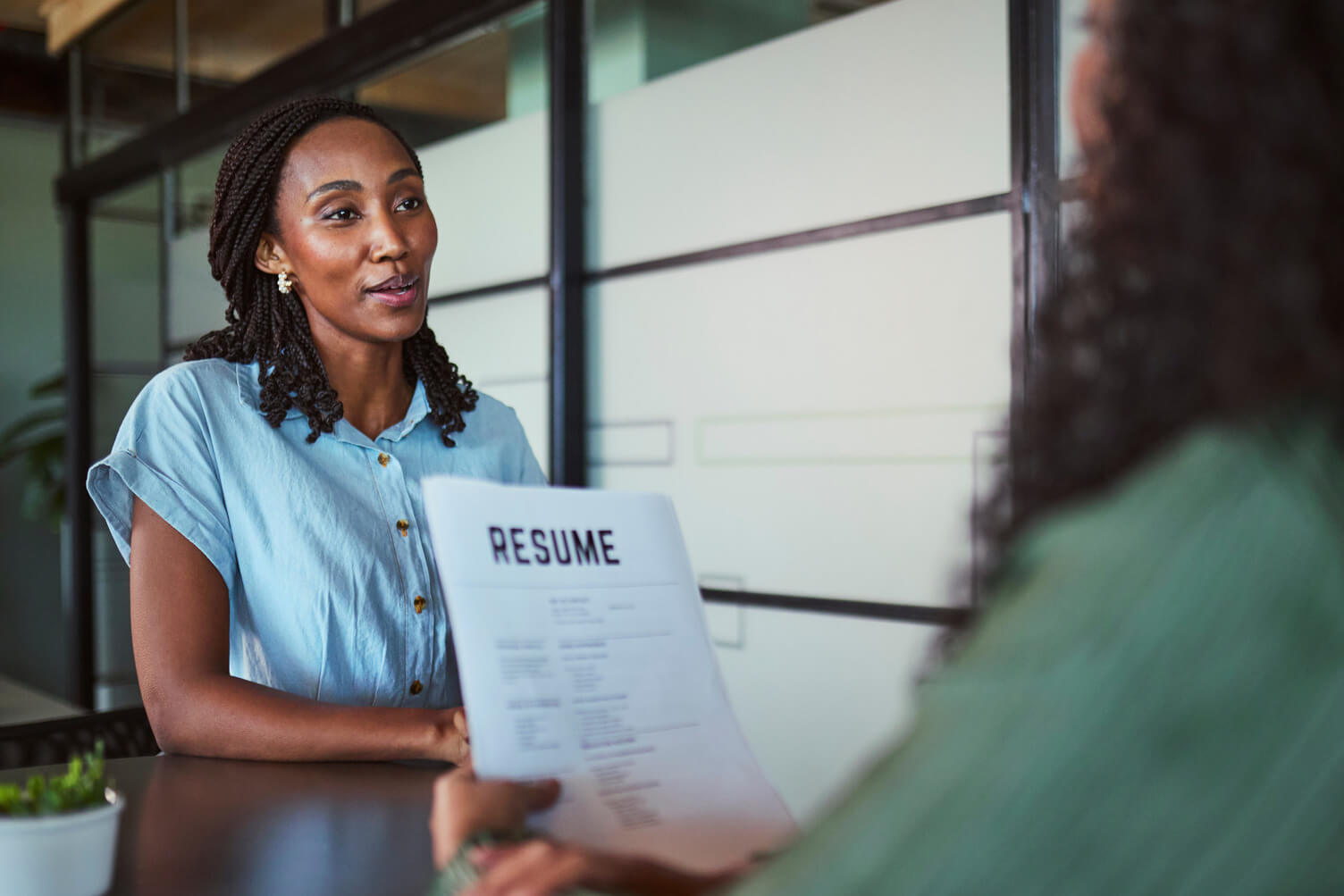 Black man in green T-shirt shakes hands with older woman across a desk in a bright office, suggesting an employment or income verification discussion.