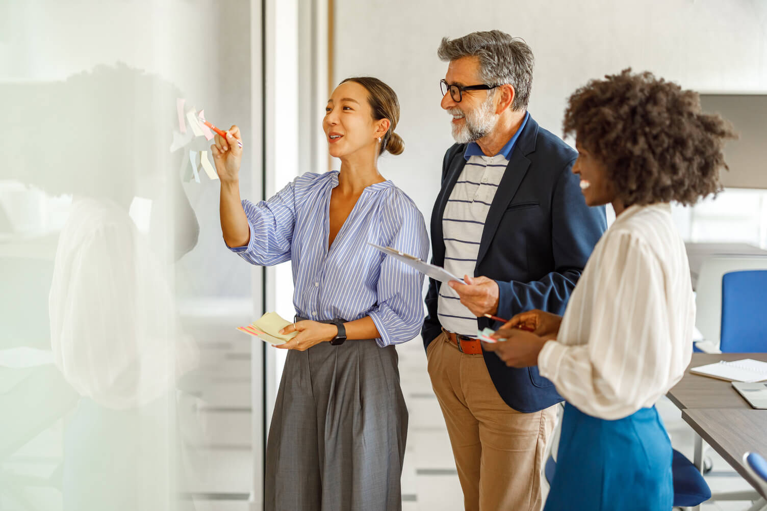Three professionals collaborate at a glass board in a bright office; a woman writes with a marker while two colleagues review notes and discuss information.