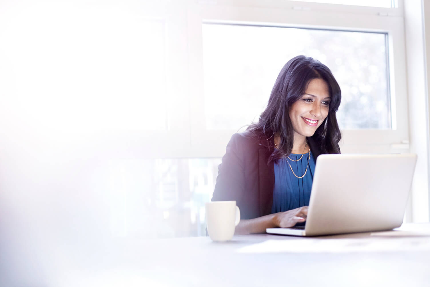 A person in business attire sits at a desk working on a laptop in a brightly lit room. Large windows behind them allow natural light to fill the space. On the desk are a white coffee mug and scattered papers, suggesting a professional and focused work environment.