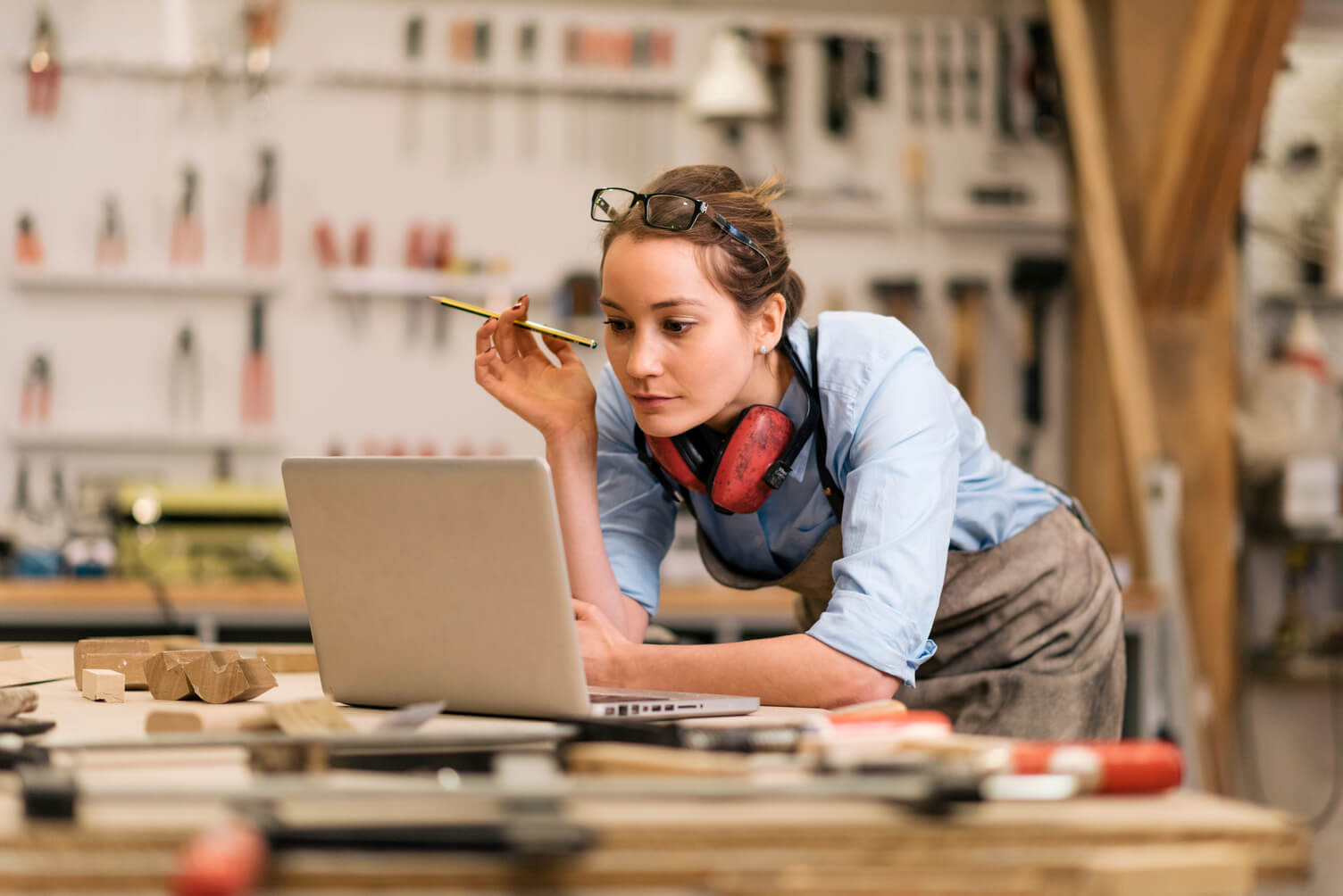 Woman in a workshop-style workspace leans over a workbench, using a laptop while holding a pencil, with tools and materials visible in the background, representing workforce management and digital employment verification through Experian Verify™ employer services.