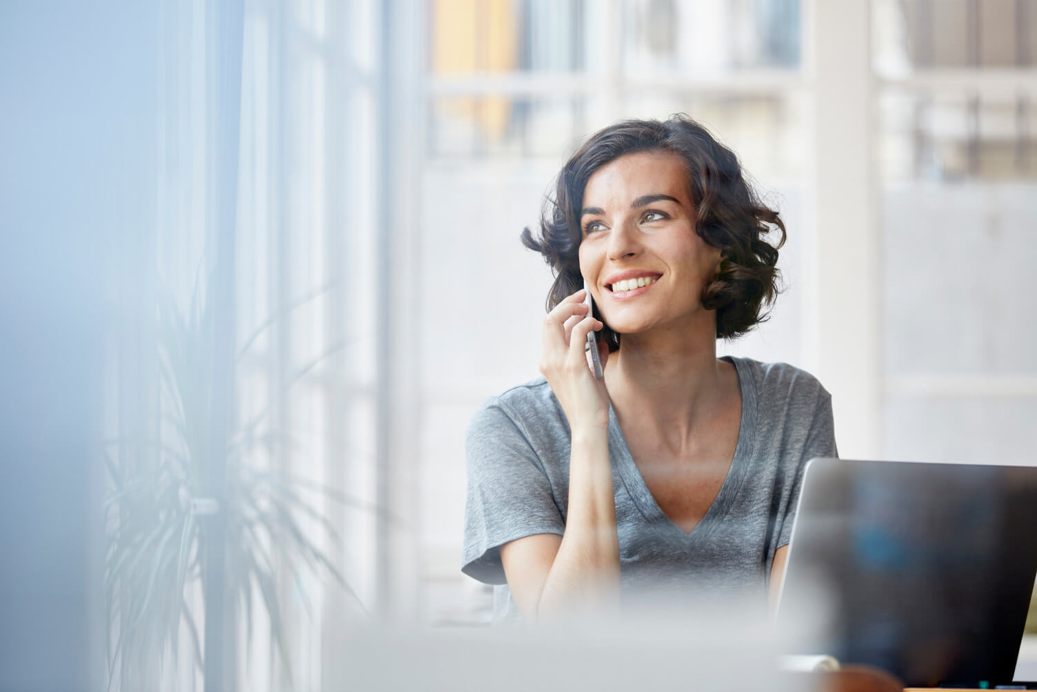 A person with short curly hair is sitting indoors near a large window, smiling while talking on a mobile phone. They are wearing a gray t-shirt and looking slightly upward. In front of them is an open laptop. The background shows a blurred view of greenery outside the window, suggesting a relaxed, casual work environment.