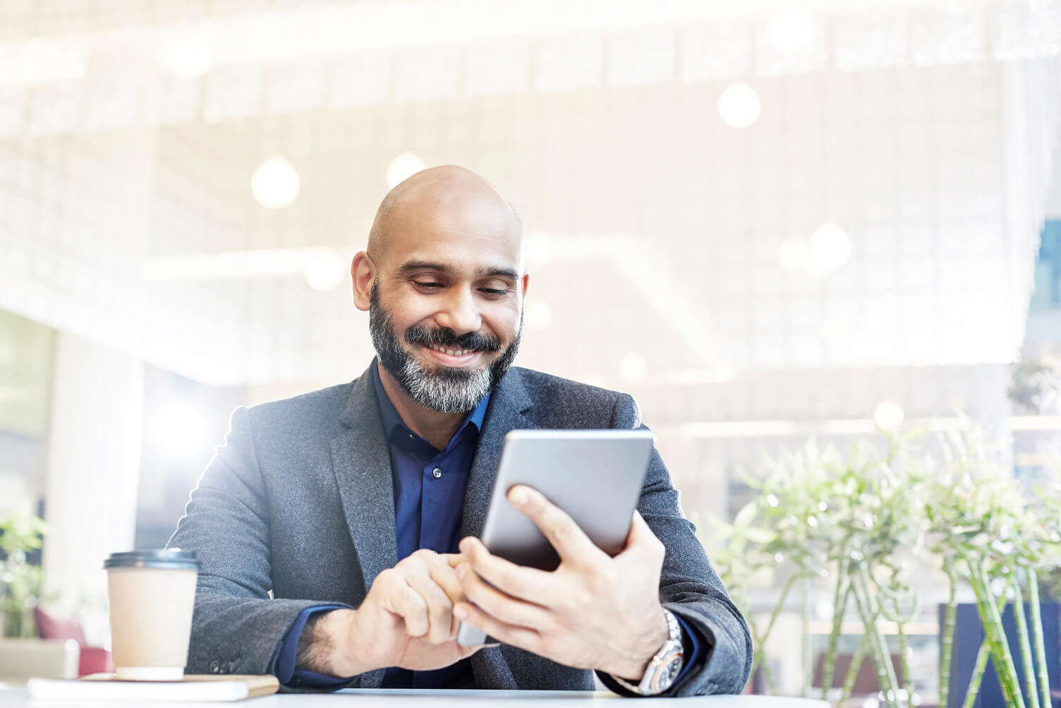 Middle-aged man with a beard in business attire sits at a table in a bright modern workspace, using a tablet with a coffee cup nearby, representing secure digital employment and income verification through Experian Verify™ employer services.