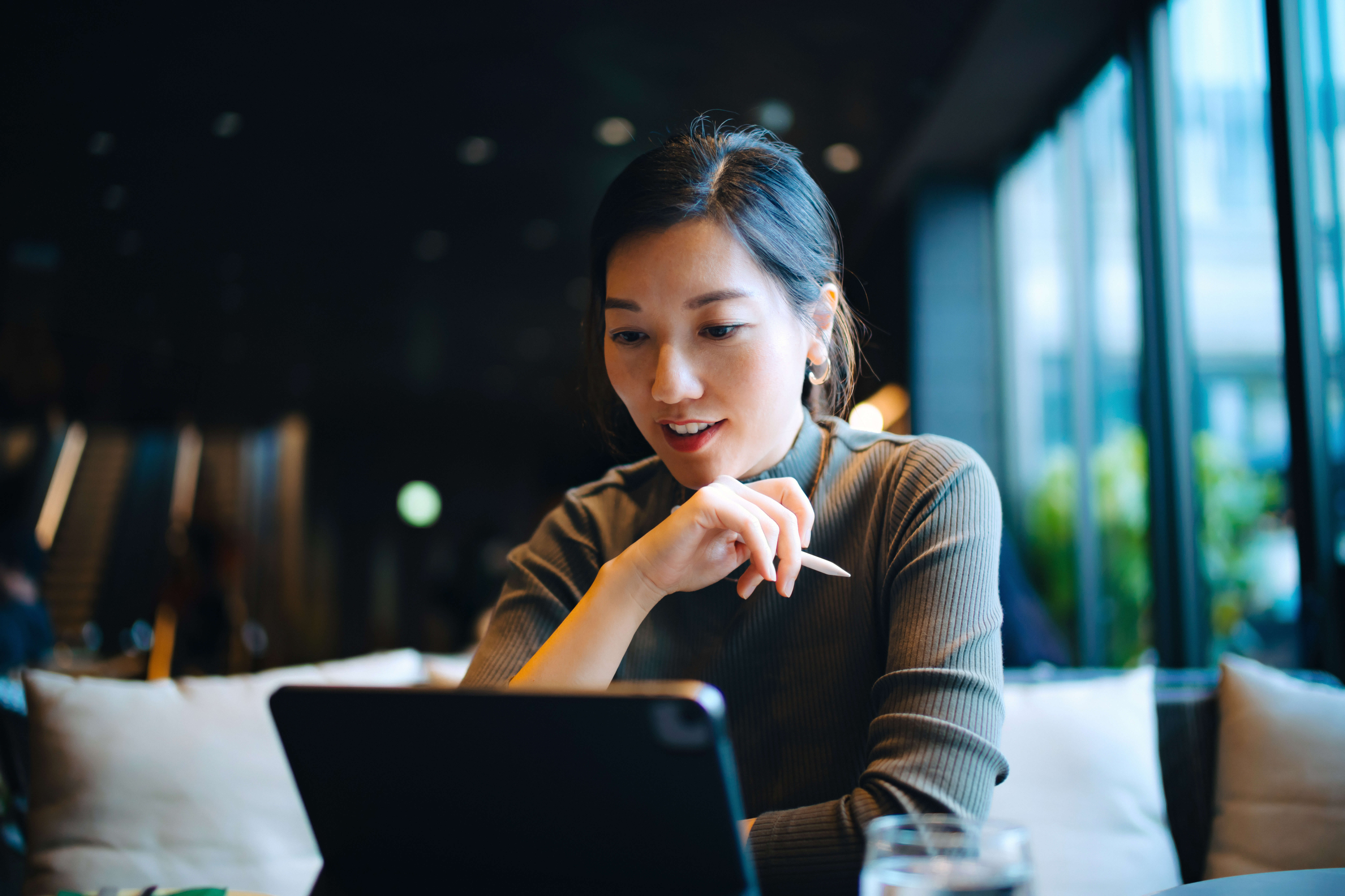 Asian woman in business-casual attire sits in a modern indoor workspace, reviewing information on a tablet, with soft lighting and blurred seating in the background, representing digital employment and income verification